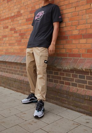 Personne portant un pantalon cargo beige, un t-shirt graphique Nike noir et des baskets noires et blanches, debout contre un mur en briques rouges en extérieur.