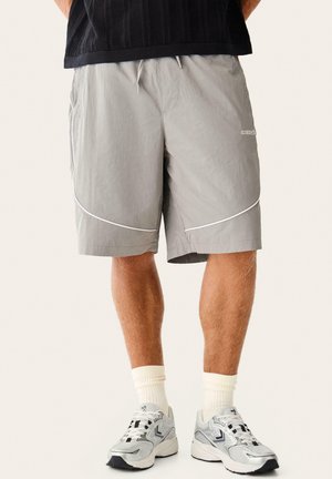 Mężczyzna w jasnoszortach sportowych z odblaskowymi paskami, białych skarpetach sportowych i srebrnych butach do biegania na neutralnym tle.