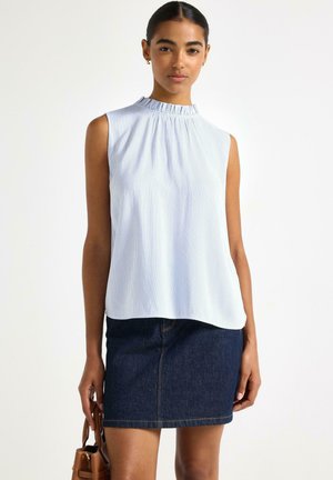 Kvinde iført en ærmeløs hvid bluse med flæset krave og en mørkeblå denimnederdel, der holder en brun håndtaske.