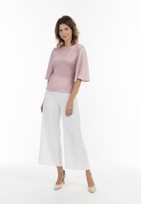 Hellrosa Bluse mit fließenden Glockenärmeln, kombiniert mit weißen weit geschnittenen Hosen. Das Outfit wird durch hautfarbene High Heels ergänzt.