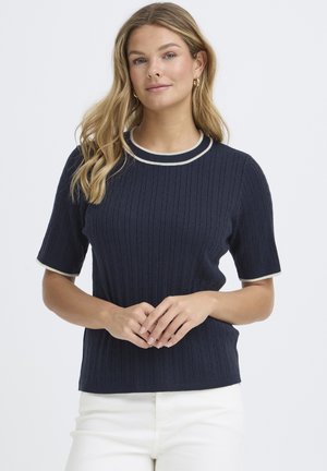 Jonge vrouw met lang blond haar, gekleed in een marineblauwe kabelgebreide trui met korte mouwen en een witte broek, staand met handen gevouwen.