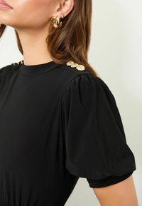 Žena v černém topu s balónovými rukávy a zlatými dekorativními knoflíky na rameni, s několika zlatými kroužkovými náušnicemi na levém uchu.