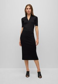 Schwarzes Kleid mit halbzip Geflecht, kurzen Ärmeln und einer Länge bis zur Mitte der Waden. Strukturiertes Gewebe mit einem subtilen Muster. Kombiniert mit schwarzen offenen Mules.