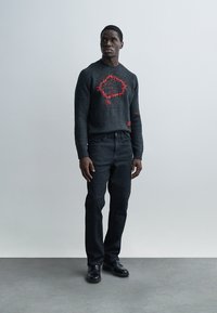 Pull gris avec motif brodé rouge, associé à un jean en denim foncé et des chaussures noires. Le mannequin se tient sur un fond uni.
