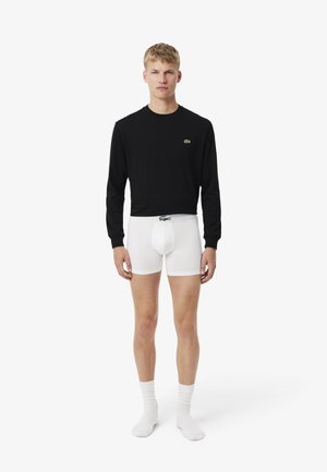 Giovane uomo in piedi, indossa boxer bianchi, maglia nera a maniche lunghe e calzini bianchi, su uno sfondo bianco semplice.