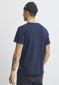 Tamsiai mėlynas trumpomis rankovėmis marškinėliai, pagaminti iš medvilnės, su apvalia apykakle, paprastas dizainas su maža etiketė ant apvado, dėvimi su tamsia mezgine kepure.