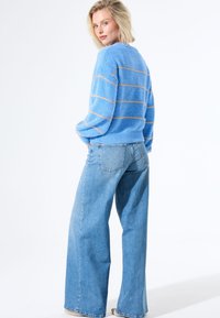 Blauer Strickpullover mit horizontalen Streifen in Hellblau und Gold, kombiniert mit hochtaillierten, hellblauen weit geschnittenen Jeans. Einfaches Texturmuster.