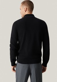 Czarny sweter dziany z prążkowanym kołnierzem i mankietami, widok od tyłu podkreślający fakturę materiału. Widoczne są szare, dopasowane spodnie.