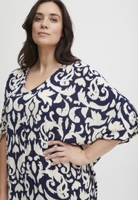 Bluza v mornarsko modri in kremni barvi s potiskom, z V-izrezom, ohlapnimi rokavi in teksturirano tkanino, ki vsebuje cvetlični vzorec.