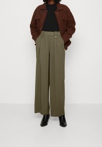 Olivenfarbene Weitbein-Hosen mit vorderen Falten und zwei Knopfdetails, kombiniert mit einem schwarzen Oberteil und einer braunen strukturierten Jacke, ergänzt durch schwarze Stiefel.