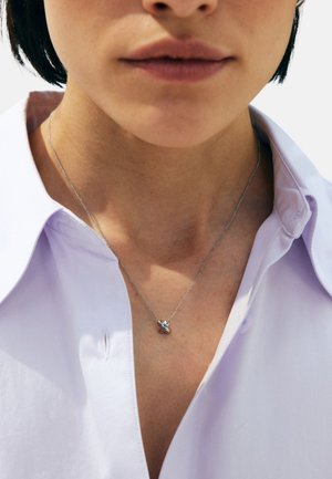 Zilveren ketting met een klein hangertje, gedragen door een persoon met een lichtpaarse blouse met kraag. Close-up beeld richt zich op de details van de ketting.