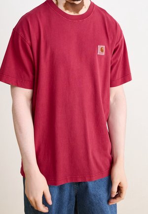 Person iført en løs rød Carhartt T-shirt med korte ærmer og blå jeans mod en ensfarvet, lys baggrund.
