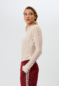 Langermet bluse med høy hals i beige blondestoff med intrikate mønstre. Kombinert med bordeaux bukser med sideaccentuer. Glatt, strukturert stoff.