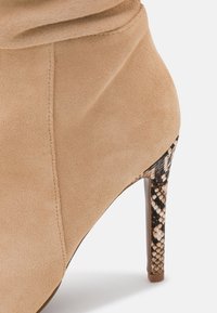 Bottine en suède beige à bout pointu et talon aiguille, avec un accent en imprimé serpent sur le talon. Texture lisse et design sans couture.