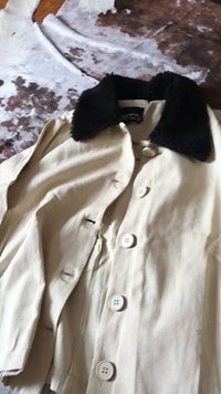 Beżowy płaszcz z dużymi białymi guzikami i czarnym sztucznym futrzanym kołnierzem, rozłożony na płasko na brązowo-białej wzorzystej powierzchni.