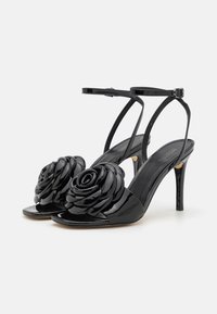 Mustakko patent leather korkokengät, joissa on kukkakuvio varpaissa ja ohut nilkkahihna. Avoin varvas ja sulavalinjainen stiletto-korko.