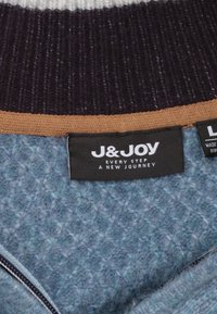 Gebreide trui met een donkerpaarse ribbelkraag, een bruine streep en een lichtblauwe gestructureerde body. Etiket leest "J&JOY" met bijgevoegde verzorgingsinstructies.