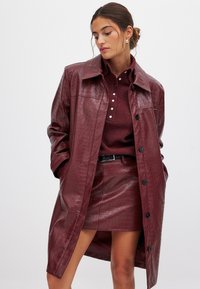 Cappotto trench in ecopelle bordeaux con texture a coccodrillo, chiusura a bottoni e colletto oversize, abbinato a una mini gonna coordinata.