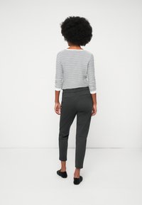 Mulher com cabelo encaracolado vestindo uma blusa de manga longa cinza e branca com riscas, calças cinza escuras e sapatos pretos sem atacadores, de pé contra uma parede branca.