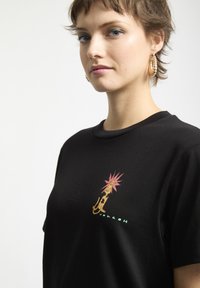 Persona joven con pelo corto que lleva una camiseta negra con un gráfico colorido de un león y pendientes de aro, con un fondo liso.