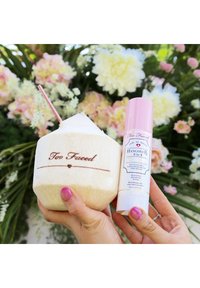 Too Faced kokosnoot-vormige container met een witte bovenkant en gegraveerd logo, naast een roze sprayfles met het label "Hangover 3-in-1."