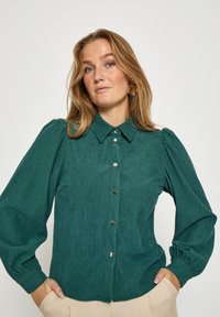 Moteris šviesiais rudais plaukais, vilkinti žalsvai mėlyną kordurovinę palaidinę su sagomis ir smėlio spalvos kelnes, stovinti su rankomis kišenėse prieš paprastą foną.