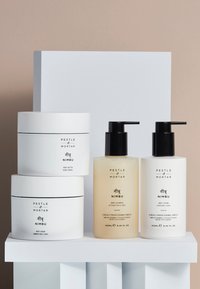 Ensemble de quatre produits de soin de la peau sur un piédestal blanc : deux flacons pompes (transparent et blanc opaque) et deux pots blancs, étiquetés avec du texte.