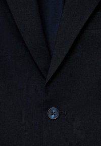 Närbild av en svart kostymjacka med slagkragar och en enda mörk knapp fäst i mitten.