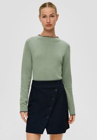 Helle grüne Langarmbluse, kombiniert mit einem dunkelblauen, gestreiften Wickelrock, der fünf schwarze Knöpfe auf der linken Seite hat.