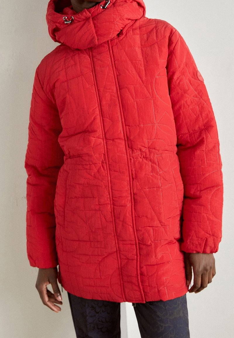 Veste rouge matelassée à capuche avec une fermeture éclair frontale portée par une personne, associée à un pantalon sombre à motifs, sur un fond uni.
