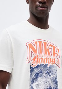 Κοντινό πλάνο ενός ατόμου που φοράει λευκό μπλουζάκι με κόκκινο και πορτοκαλί γραφικό "Nike Hoops" πάνω από σχέδιο μπλε κάγκελου αλυσίδας.