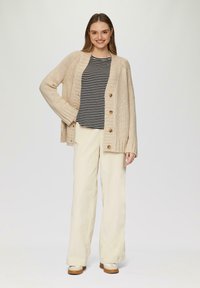Beige Strickjacke mit Holzknöpfen, getragen über einem schwarz-weißen gestreiften Shirt. Kombiniert mit hellen, weit geschnittenen Hosen und beigen Schuhen.