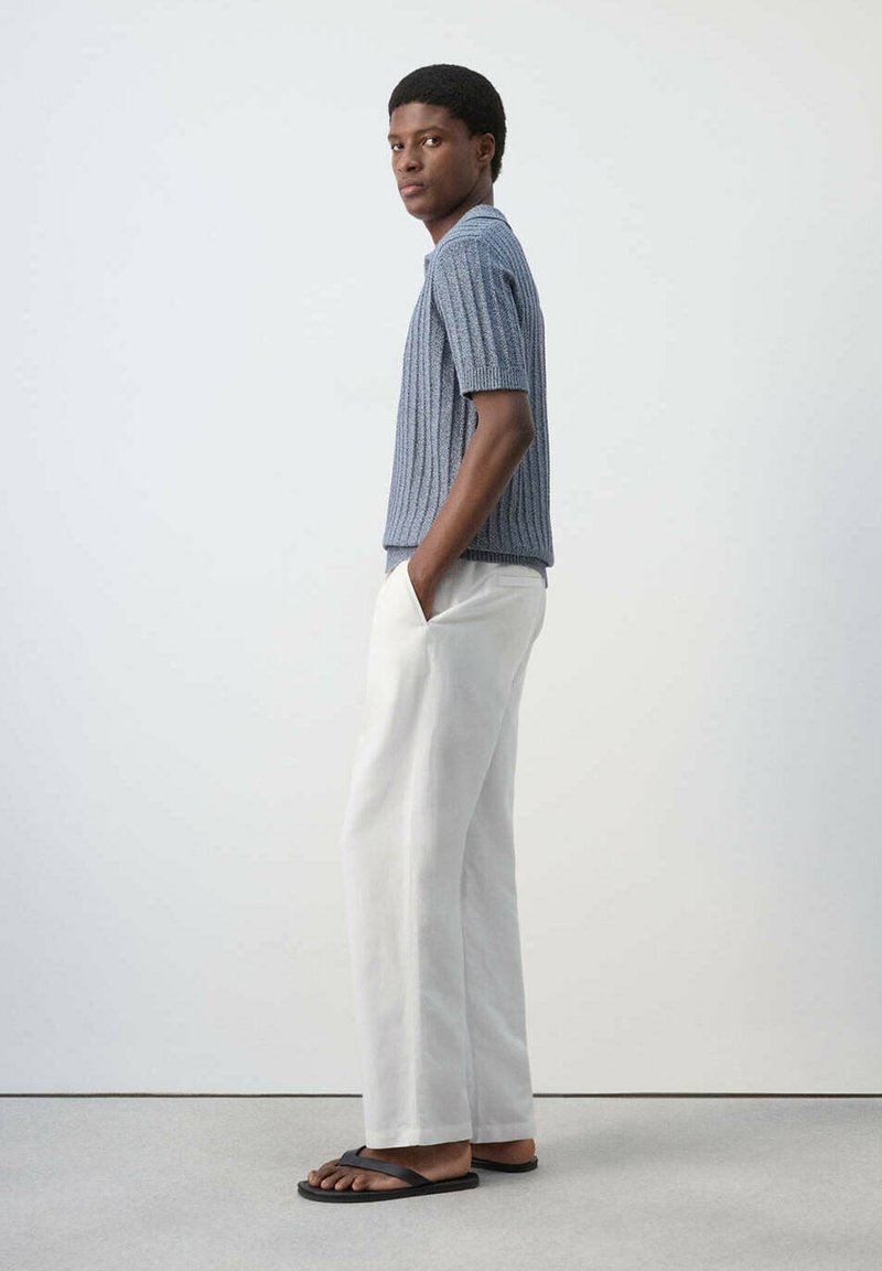 Jeune homme debout de profil, les mains dans les poches, portant un t-shirt en maille bleu à manches courtes, un pantalon blanc à jambes larges et des sandales noires.