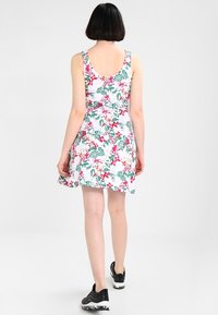 Blommönstrad klänning med vit bakgrund, livfulla gröna blad och rosa blommor. Ärmlös design med en utsvängd kjol. Svarta sneakers bärs.