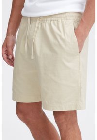 Beige cotton shorts with a drawstring waist, side pockets, and a subtle grid pattern. Hem ends just above the knee.