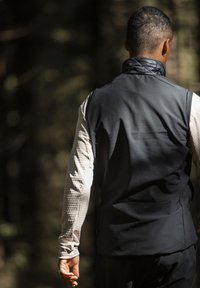 Schwarze ärmellose Jacke mit hohem Kragen über einem hellen Langarmoberteil mit strukturiertem Muster. Von hinten von einer Person getragen.