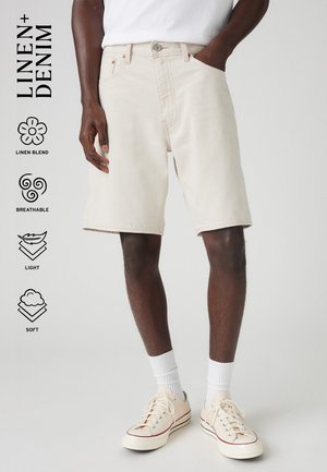 Mandlig model iført lyse beige linnedblendede denimshorts, hvide sokker og cremefarvede lærredssneakers, stående med den ene hånd i lommen.