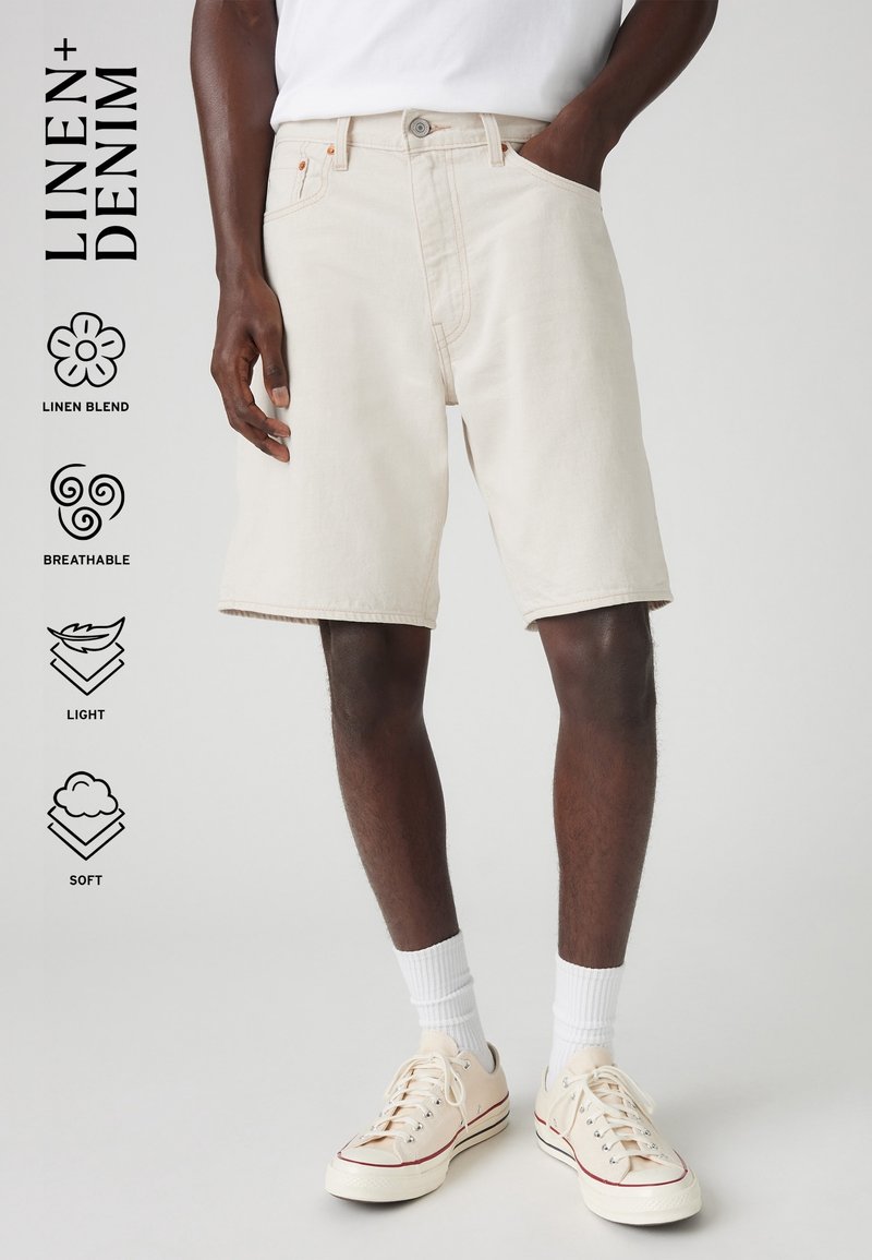 Modèle masculin portant un short en denim mélangé lin beige clair, des chaussettes blanches et des baskets en toile crème, debout avec une main dans la poche.