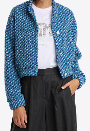Woman wearing blue textured buttoned jacket, white graphic t-shirt, and dark gray pinstripe high-waisted pants, one hand in pocket.