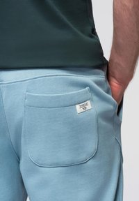 Los pantalones deportivos de un azul claro presentan un bolsillo trasero con una pequeña etiqueta de la marca. La tela es suave y ligeramente texturizada, con un diseño de corte relajado.