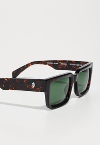 Black and tortoiseshell sunglasses with square green lenses. Thick, glossy frame featuring a logo detail on the side. No visible patterns.