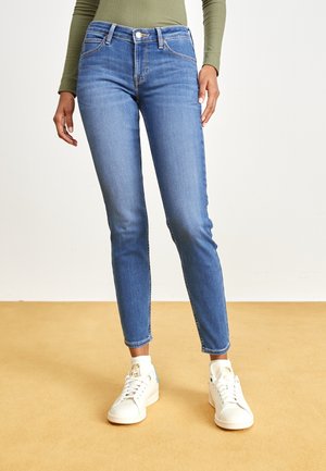 Jeans de denim azul ajustados con un aspecto deslavado, cinco bolsillos y una cintura alta. Combinados con zapatillas blancas con acentos azules.