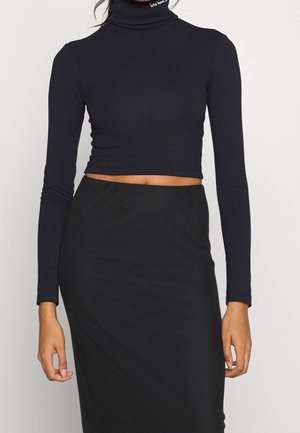 Vrouw draagt een zwart cropped coltrui met lange mouwen en een hooggesneden zwarte rok, waarbij haar buik zichtbaar is.