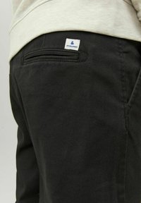 Nära bild av person som bär svarta byxor med en bakficka och vit etikett med "JACK & JONES"-logotypen.