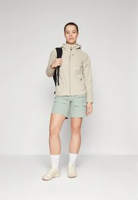 Νεαρή γυναίκα που στέκεται φορώντας ένα μπεζ ζακέτα με φερμουάρ, φωτεινά πράσινα cargo σορτς, λευκές κάλτσες, μπεζ sneakers και μια μαύρη τσάντα πλάτης στον έναν ώμο.