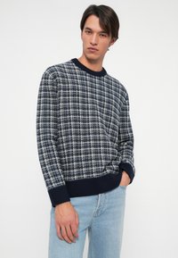 Νεαρός άνδρας με σκούρα μαλλιά, φορώντας ένα ναυτικό μπλε και λευκό καρό πουλόβερ και ανοιχτού μπλε τζιν, με το ένα χέρι στην τσέπη.