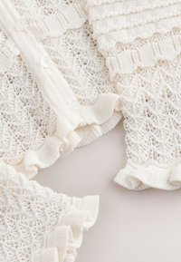 Tissu tricoté blanc avec des motifs de dentelle complexes, des bords froncés et de petits boutons blancs le long d'un vêtement plié.