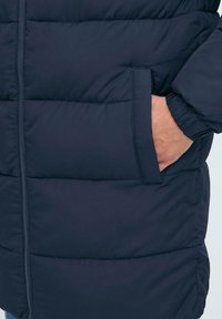 Marineblå puffjakke med sydde paneler, glidelås og sidelommer. Har elastiske mansjetter for varme. Glatt, quiltet tekstur.
