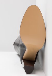 Semelle et talon d'une botte à talon haut en cuir noir vue par le dessous, avec une semelle extérieure lisse couleur tan et une fermeture éclair latérale.