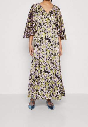 Mujer con un vestido largo de flores con hojas moradas y verdes, escote en V, mangas anchas tres cuartos y zapatos azules de punta estrecha con tiras.